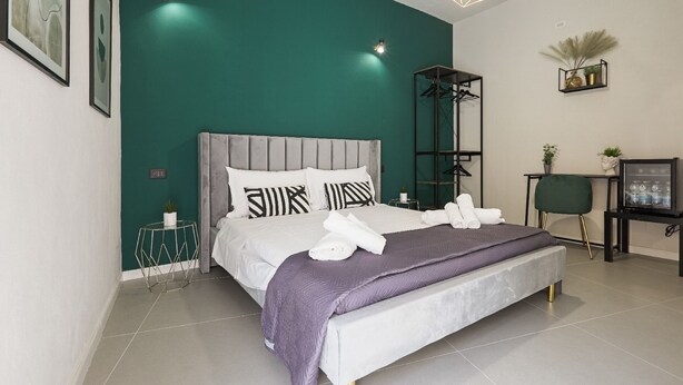 A modern bedroom with a grey upholstered bed, a purple throw blanket, and neatly folded white towels. - Chianu Palì guest rooms (Agrigento)