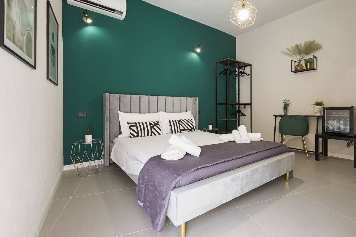 A modern bedroom with a grey upholstered bed, a purple throw blanket, and neatly folded white towels.