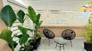 A covered patio area with a tiled floor, two black wireframe chairs, a small glass table, and large potted plants. - Chianu Palì guest rooms (Agrigento)