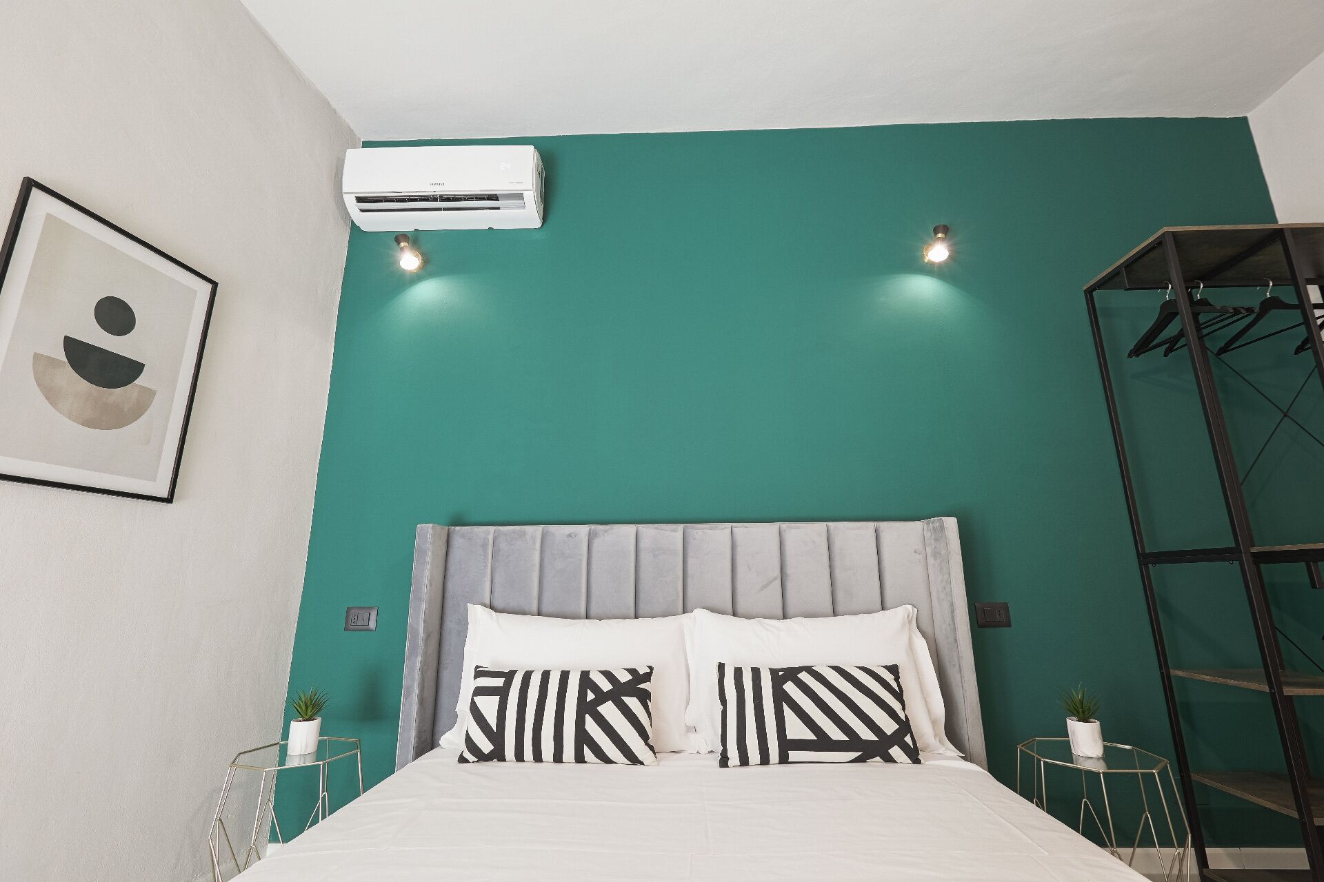 A bedroom with teal walls, a grey upholstered headboard, and a bed with black and white striped pillows. There is a white air conditioning unit mounted on the wall, and a framed artwork above the bed.