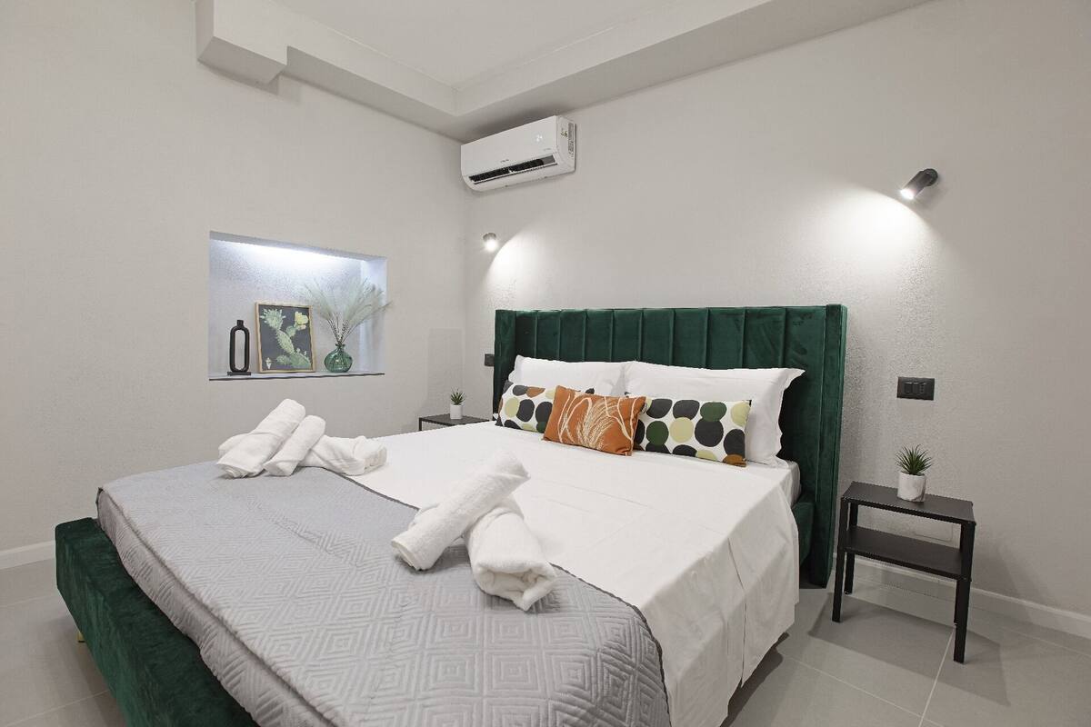 A neatly made bed with white linens and a green headboard, a small bedside table with a plant, and a wall-mounted air conditioner.