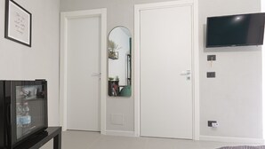 A modern living room with a flat-screen TV, a glass-fronted refrigerator, a mirror, and a door. - Chianu Palì guest rooms (Agrigento)