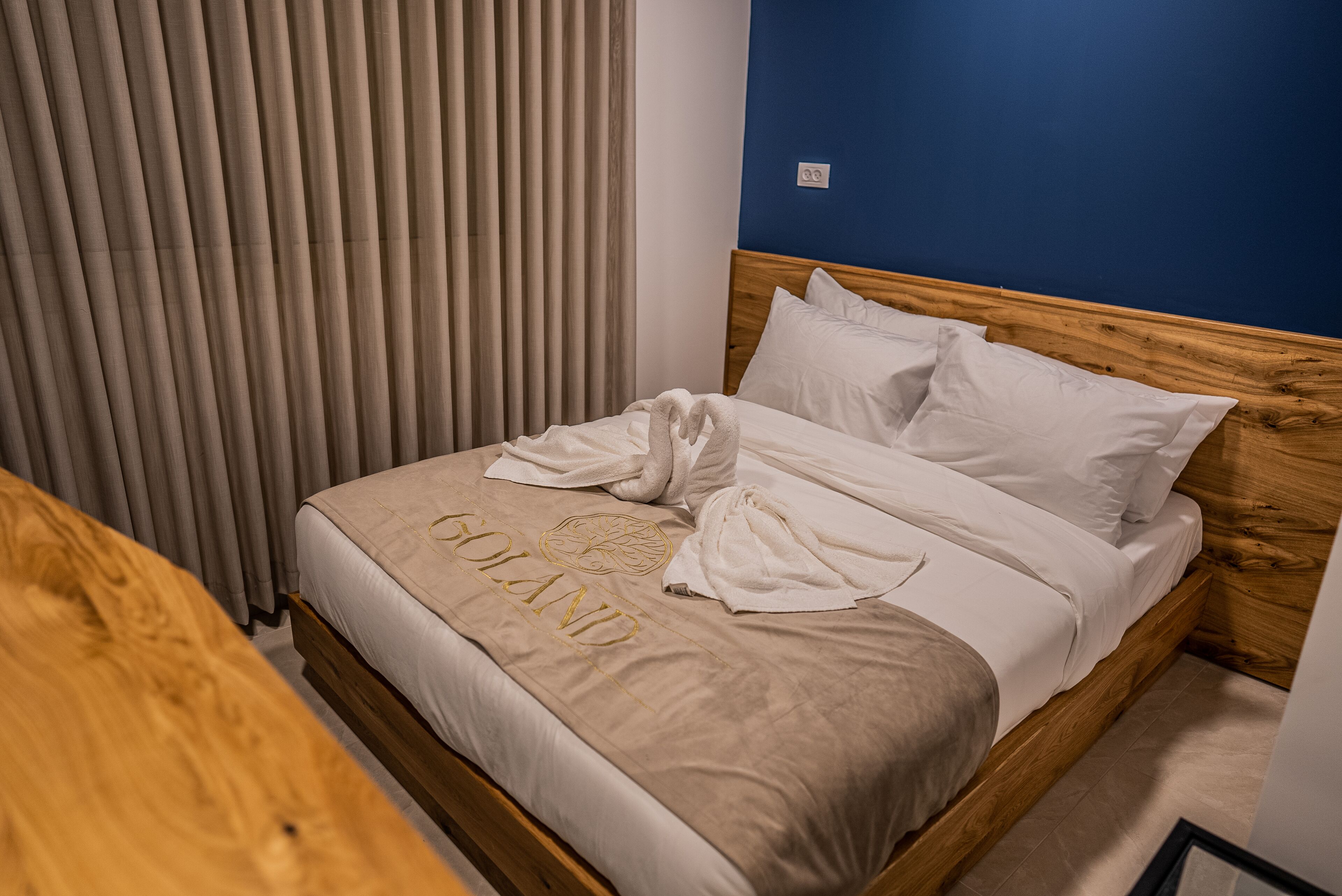 Una cama bien hecha con cabecera de madera y ropa de cama blanca, una mesita de noche de madera y una ventana con persianas verticales.