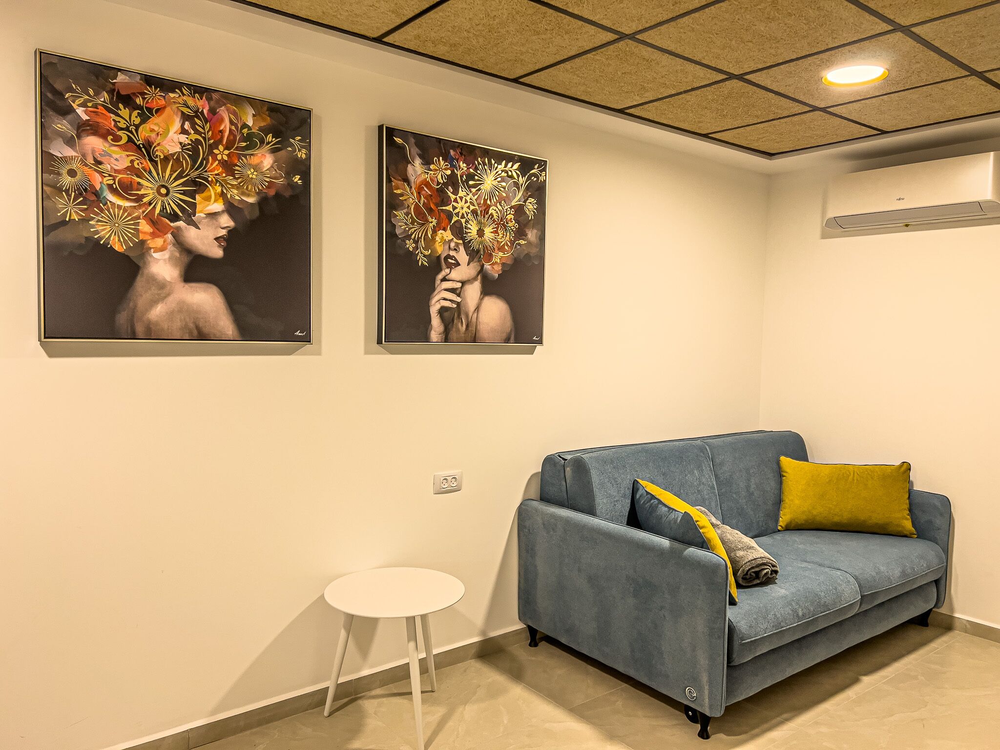 A modern living room with a blue sofa, a small white stool, and two framed artworks on the wall.