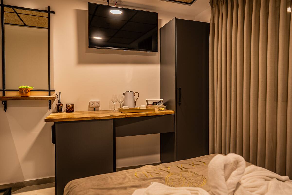 A bedroom with a bed, a desk, a mirror, a TV, and a window with curtains.
