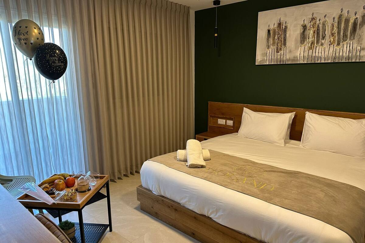 A hotel room with a large bed, a wooden headboard, white bedding, and a breakfast tray with fruits and a glass.