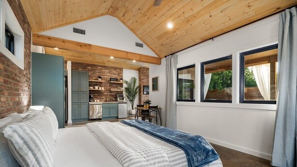 A bedroom with a large bed, a wooden ceiling, a brick wall, and a view of a kitchen and dining area. - Hot tub, Fireplace, Studio, Close to Main St. (Fredericksburg)