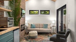 A cozy room with a brick wall, a sofa, a small ottoman, a chair, a kitchen area with a refrigerator, and a ceiling fan. - Hot tub, Fireplace, Studio, Close to Main St. (Fredericksburg)