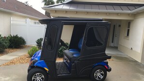 A compact, blue three-wheeled vehicle parked in front of a house with a white fence and a covered garage. - 1194 Flamingo Pl (The Villages)
