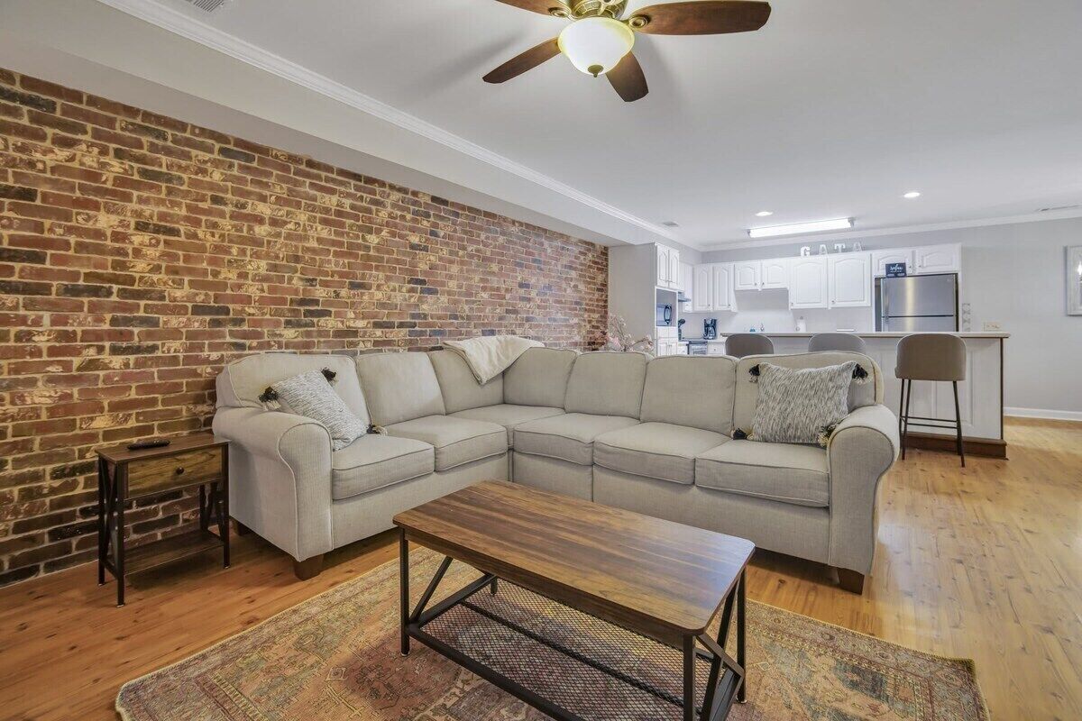 Ein Wohnzimmer mit Ecksofa, Holztisch, Ziegelwand und Deckenventilator.