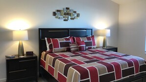 Een net opgemaakt bed met een rood-beige sprei, twee bedlampjes, een digitale klok en een kunstwerk boven het bed.