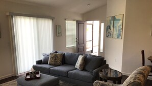 Een woonkamer met een grijze bank, een gestreept voetenbankje en een fauteuil, een klein bijzettafeltje en een groot raam met jaloezieën.