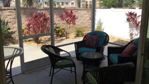 Un patio cubierto con mobiliario de mimbre, una mesa de vidrio y vistas a un barrio residencial con palmeras y casas.