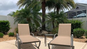 Två beige fåtöljer med metallstativ och ett litet bord, placerade på en betongterrass med en palm och olika buskar i bakgrunden.