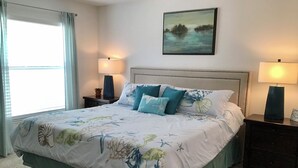 Une chambre à coucher avec un lit, deux tables de chevet, une commode, une fenêtre avec des rideaux et un tableau au mur.