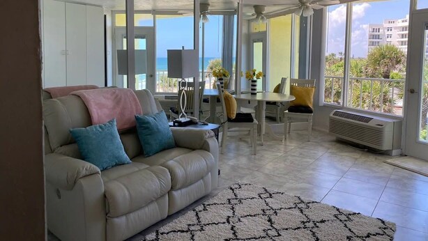 Un salon comprenant un canapé, une table à manger et des chaises. Une grande fenêtre offre une vue sur la mer.