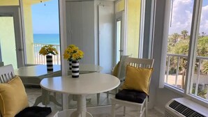 Une pièce avec une table et des chaises blanches, un vase rempli de fleurs jaunes et une vue sur l’océan.