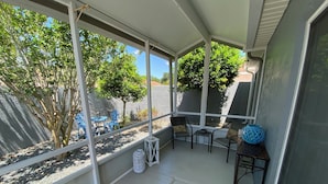 Un porche cubierto con una zona de estar, que incluye sillas y una mesa, con vista a un jardín con árboles y otra mesa de patio.