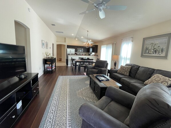 A living room with a television, sofa, coffee table, and a rug. There is a dining area with chairs and a table, and a kitchen in the background. - 9238 SE 166th Sprung Ln (The Villages)