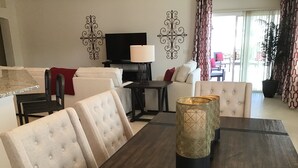Una sala moderna con una mesa de comedor de madera oscura, sillas blancas acolchadas, un televisor de pantalla plana y cuadros decorativos en la pared.