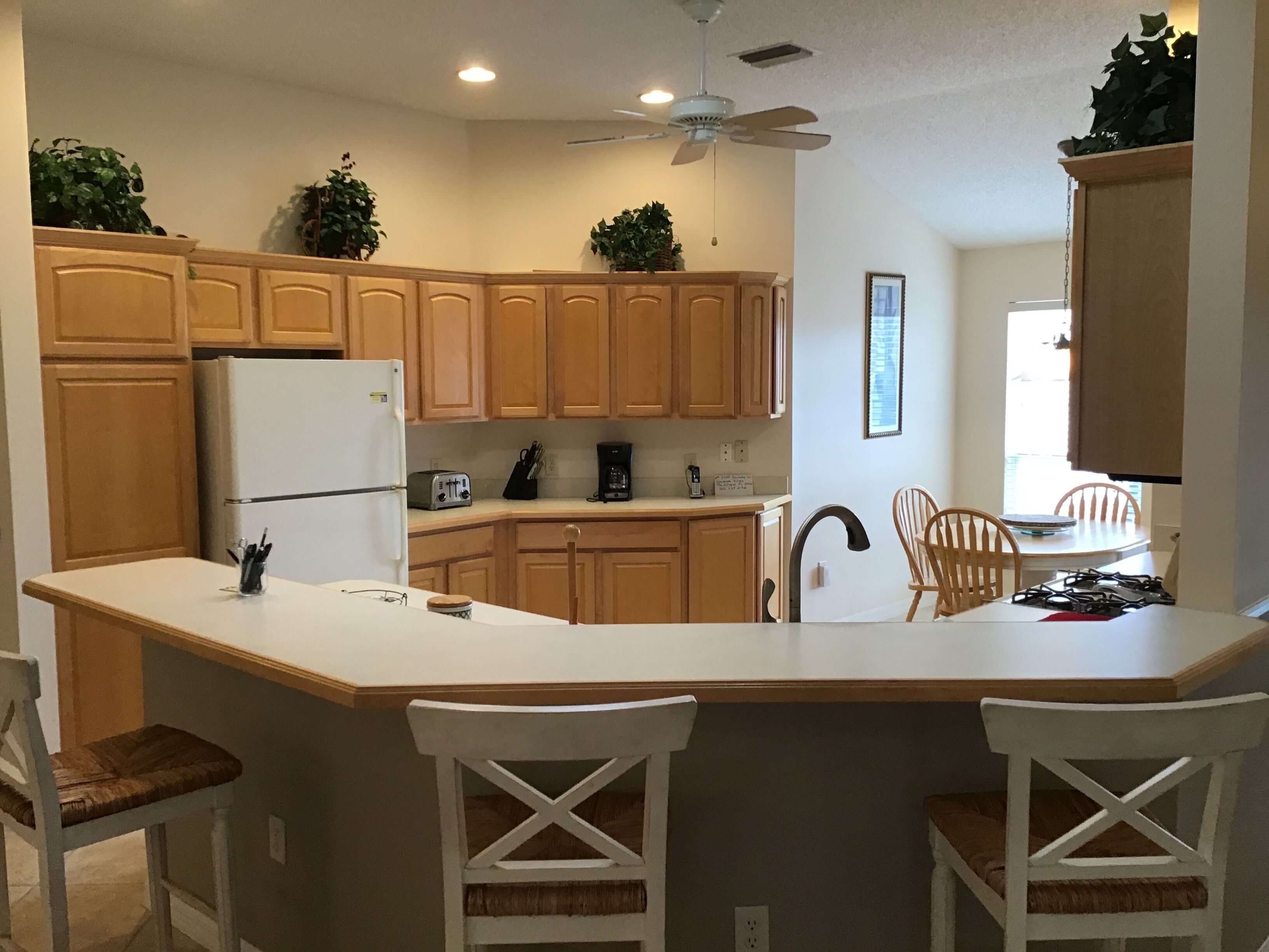 A kitchen with wooden cabinets, a white refrigerator, a built-in oven, a coffee maker, and a ceiling fan. There are two white bar stools and a dining table with chairs.