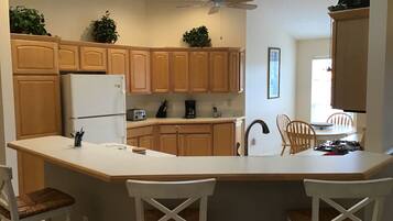A kitchen with wooden cabinets, a white refrigerator, a built-in oven, a coffee maker, and a ceiling fan. There are two white bar stools and a dining table with chairs.