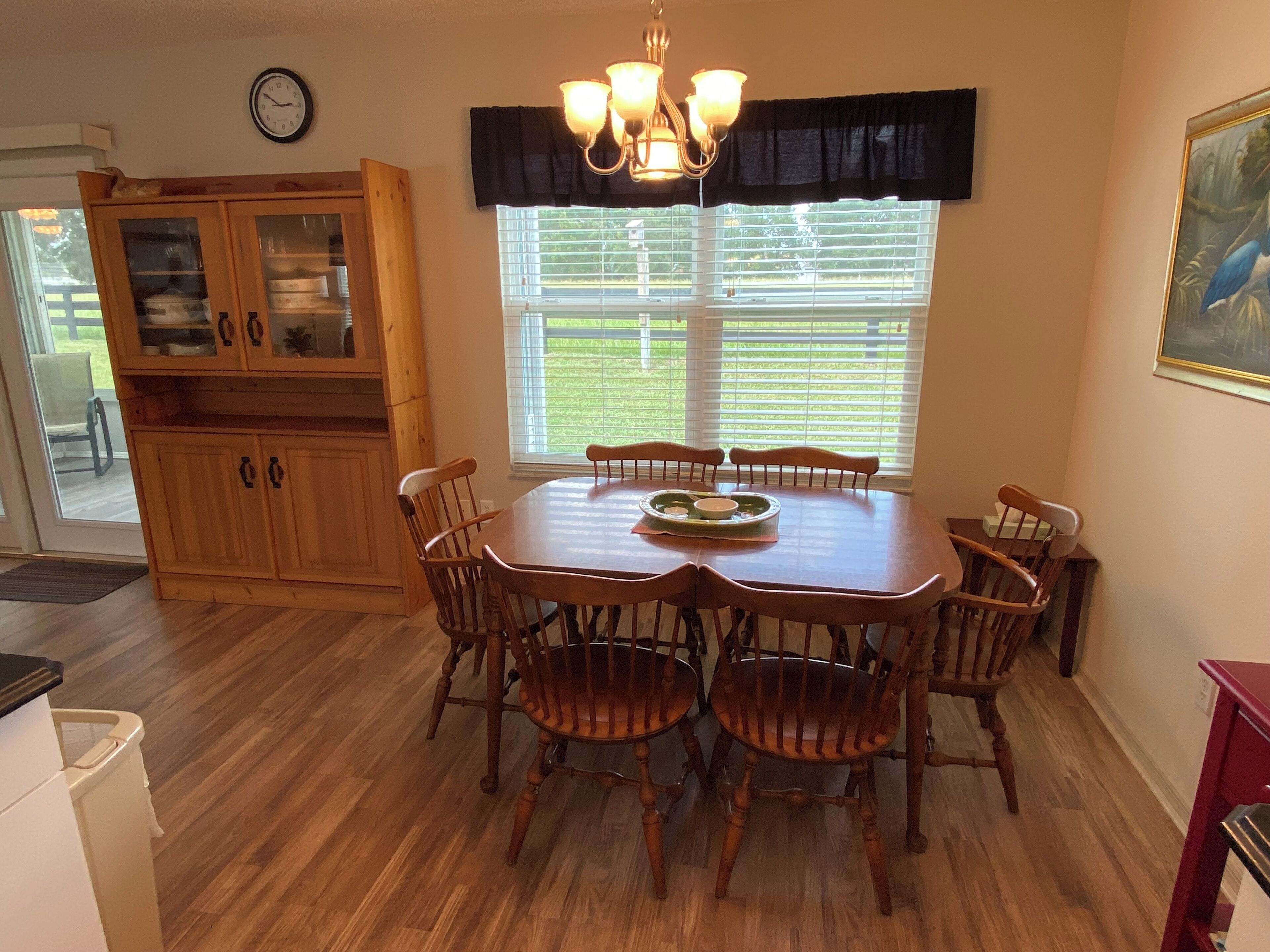 A dining area with a wooden table and chairs, a chandelier, and a window with blinds.