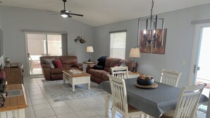 A spacious living room with a dining area, featuring a ceiling fan, a chandelier, and a large window with blinds. - 2066 Duval Ct (The Villages)