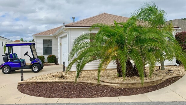 A residential house with a white exterior and a brown roof, a small garden with a palm tree, and a blue golf cart parked in front. - 2066 Duval Ct (The Villages)