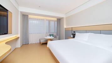 A hotel room with a large bed, a bedside table, a small round table with a chair, a TV mounted on the wall, and a window with sheer curtains.
