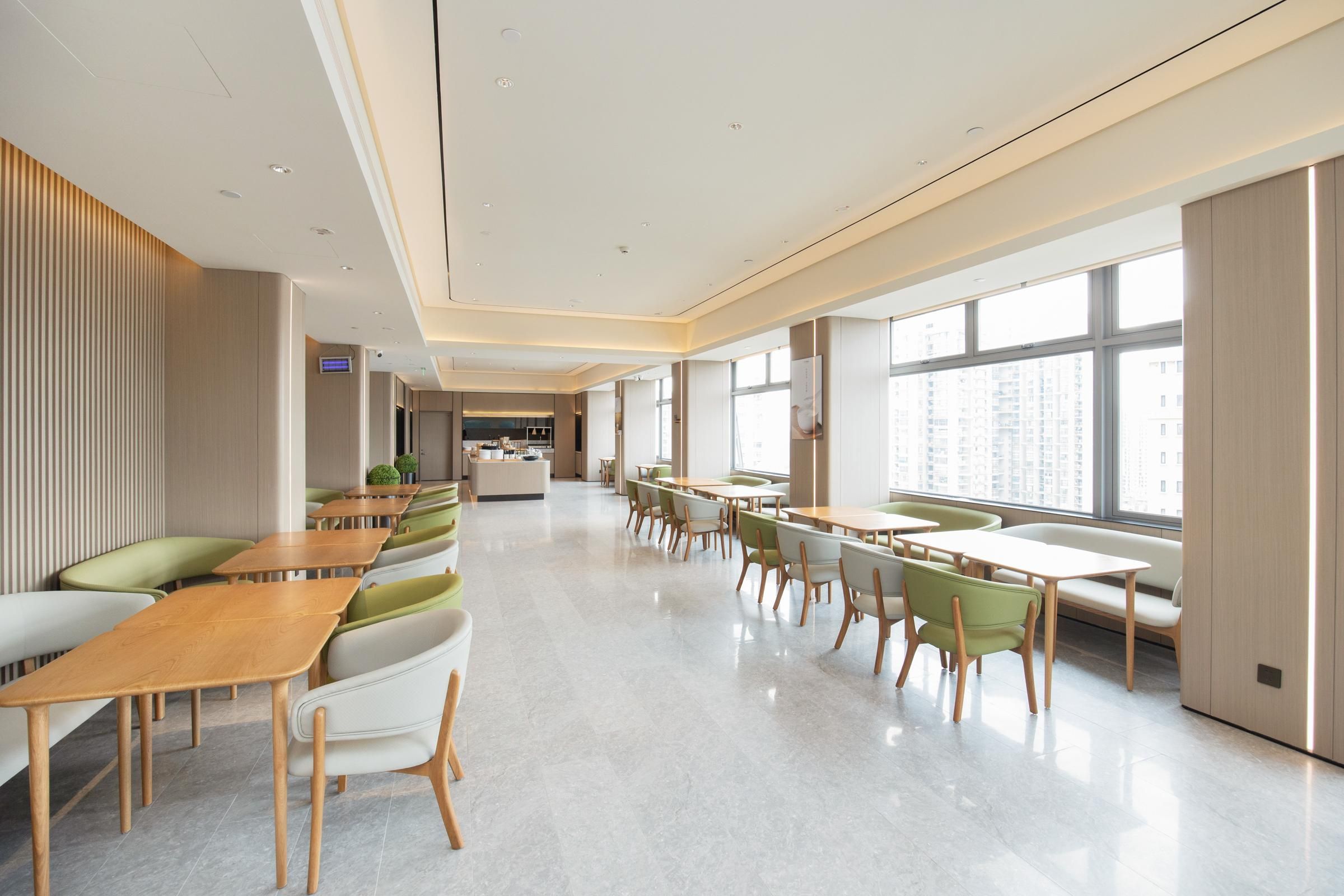 A modern hotel lobby with a long reception desk, seating areas with green and white chairs, and large windows offering a view of the city.