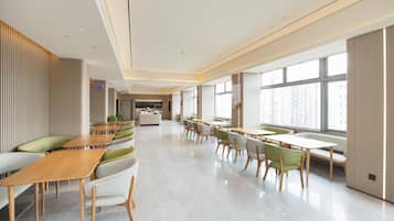 A modern hotel lobby with a long reception desk, seating areas with green and white chairs, and large windows offering a view of the city.