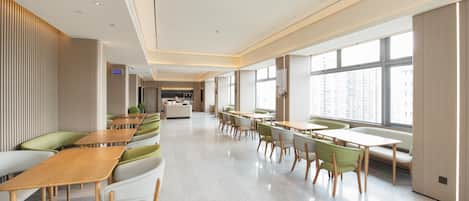 A modern hotel lobby with a long reception desk, seating areas with green and white chairs, and large windows offering a view of the city.