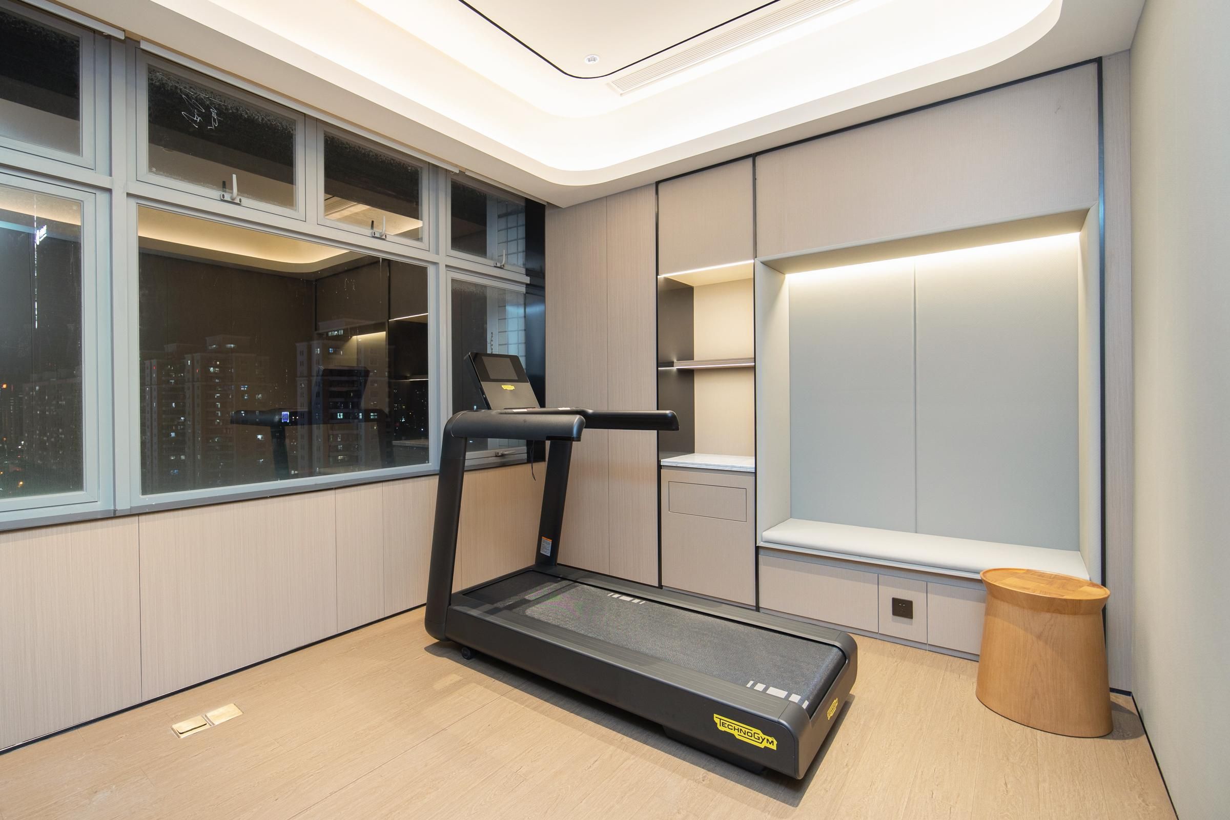 Una habitación moderna con una cinta de correr, una ventana con vista a la ciudad, estanterías empotradas y un pequeño taburete de madera.