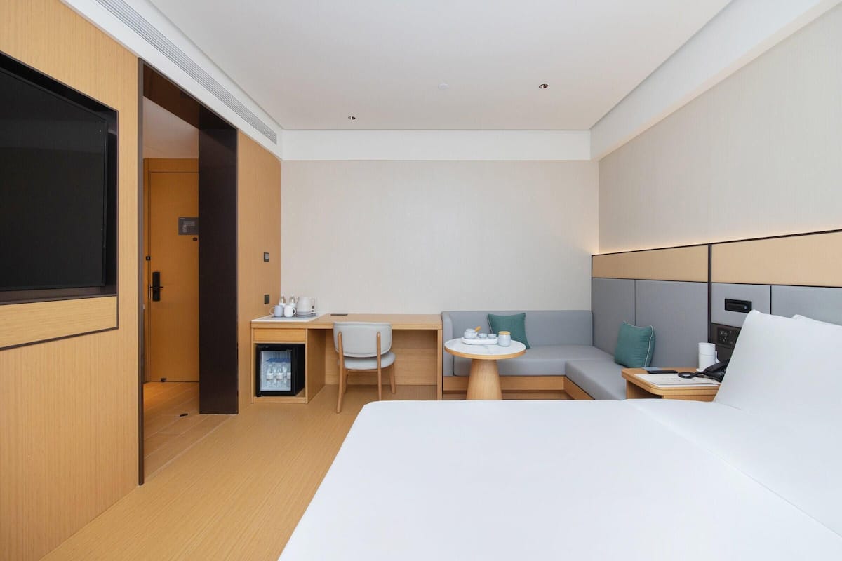 A modern hotel room with a bed, a desk with a chair, a sofa, and a small table with a lamp.