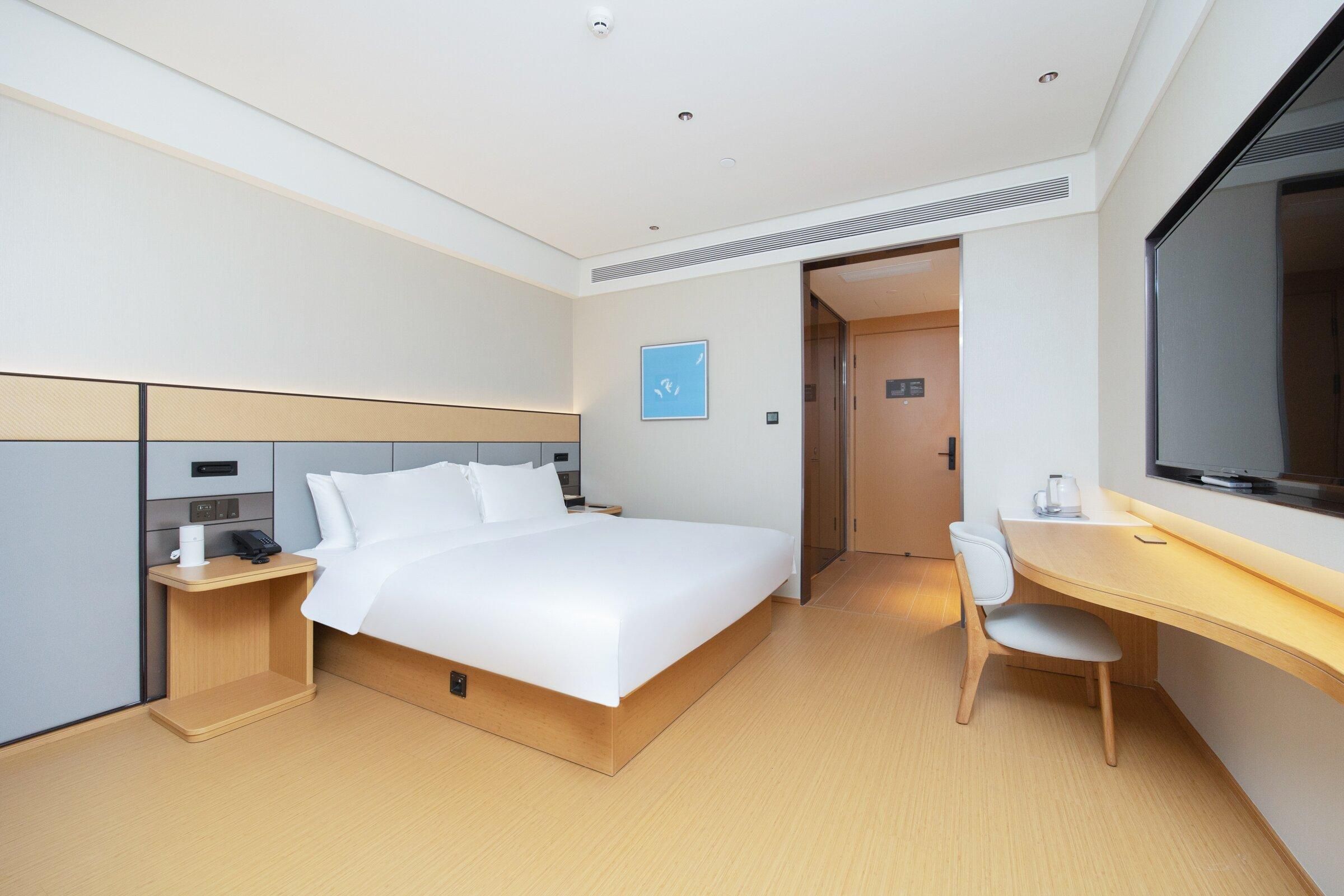 A modern hotel room with a bed, bedside table, chair, desk, and a large window.