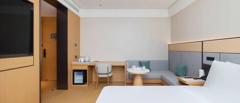 A modern hotel room with a bed, a desk with a chair, a sofa, and a small table with a lamp.