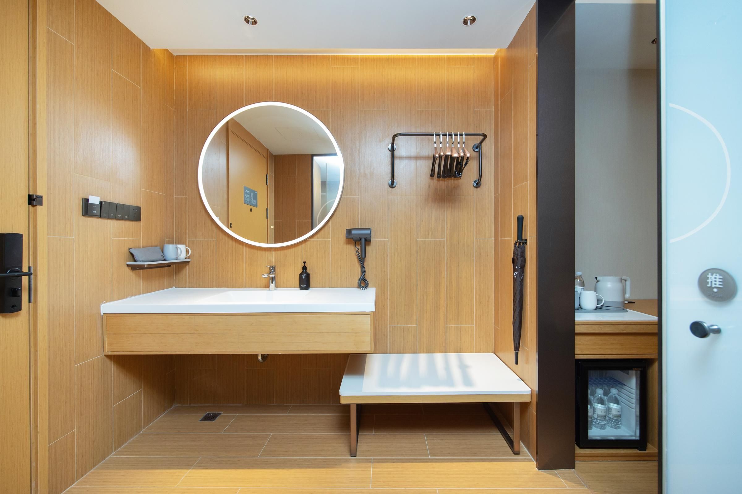 Un baño moderno con tocador de madera, un espejo redondo, un perchero para toallas y un refrigerador pequeño.
