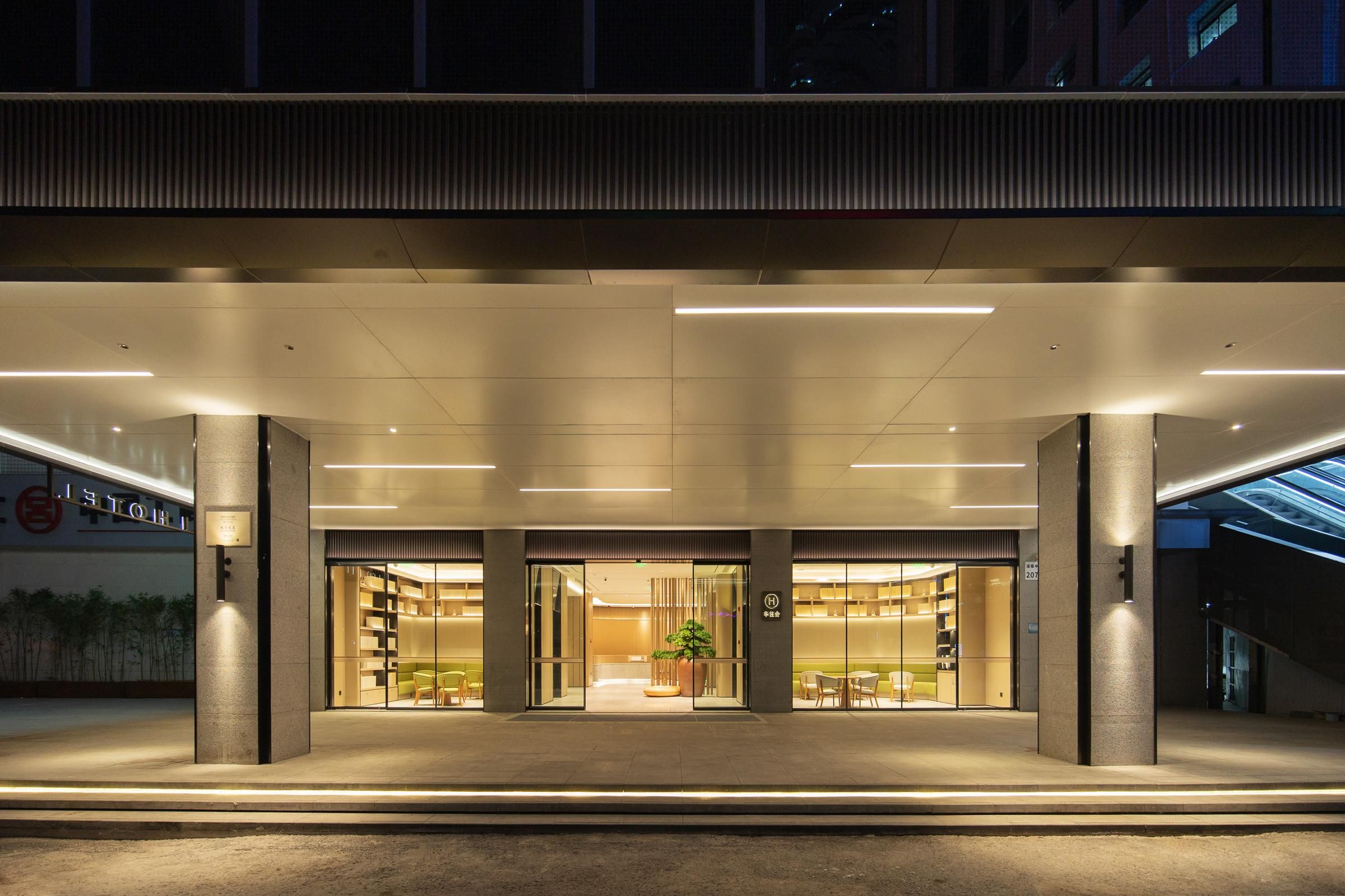 Una entrada de hotel moderna con fachada de vidrio, una planta central y zonas de estar visibles a través de las puertas.