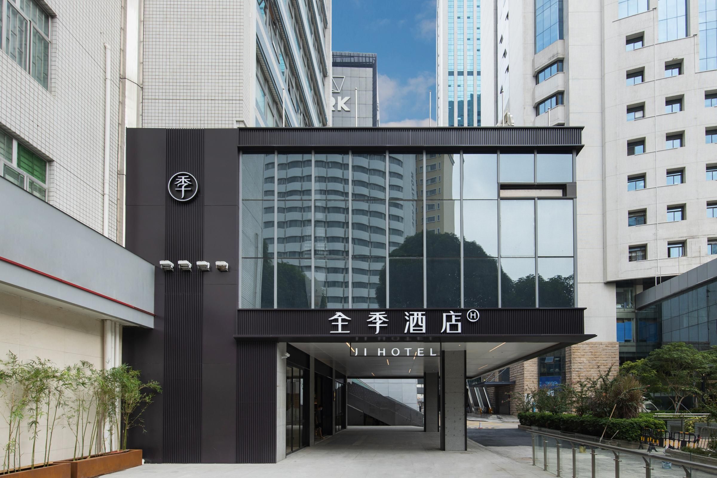 Entrada de un hotel moderno con fachada de vidrio, un letrero con caracteres chinos y un cielo despejado al fondo.