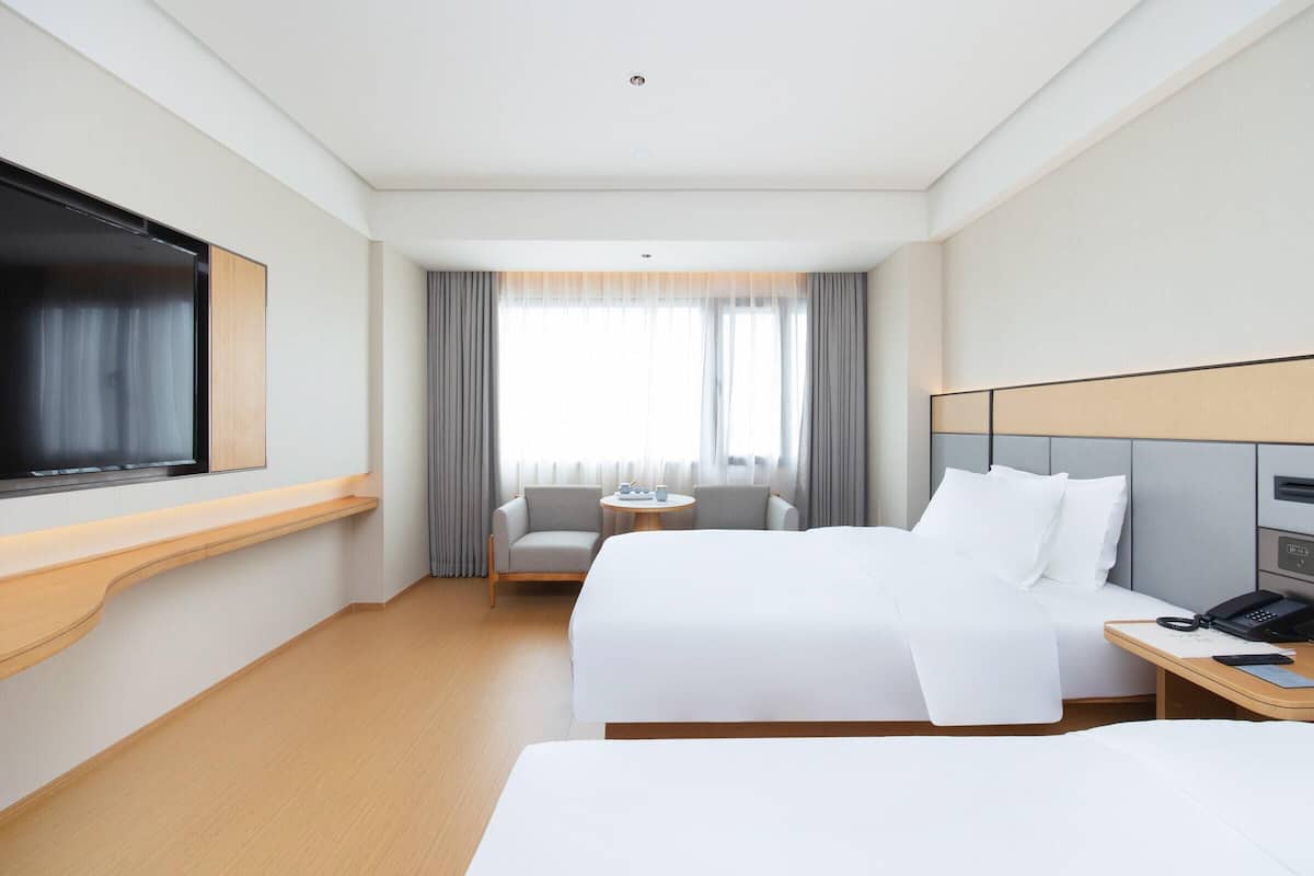 A hotel room with a bed, a TV, a desk with a phone, a chair, and a window with curtains.