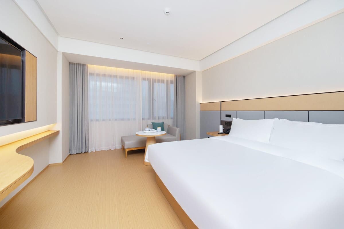 A hotel room with a large bed, a bedside table, a small round table with a chair, a TV mounted on the wall, and a window with sheer curtains.