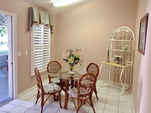 A dining area with a round glass table, four wicker chairs with floral cushions, a vase with flowers, and a metal shelf with decorative items.