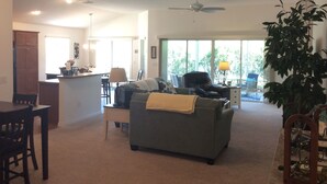 A spacious living room with a grey sofa, a dining area with a table and chairs, a kitchen in the background, and a ceiling fan. - 2067 Useppa Oaks Ln (Wildwood)