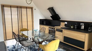 Une cuisine moderne avec une table à manger en verre, des chaises noires et une chaise jaune. On y trouve une cuisinière noire, une machine à café et un four à micro-ondes sur le plan de travail.
