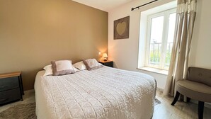 A neatly made bed with a quilted cover, a bedside lamp, and a chair near a window with curtains. - BIG HOUSE N°6 (Ferrières-la-Verrerie)