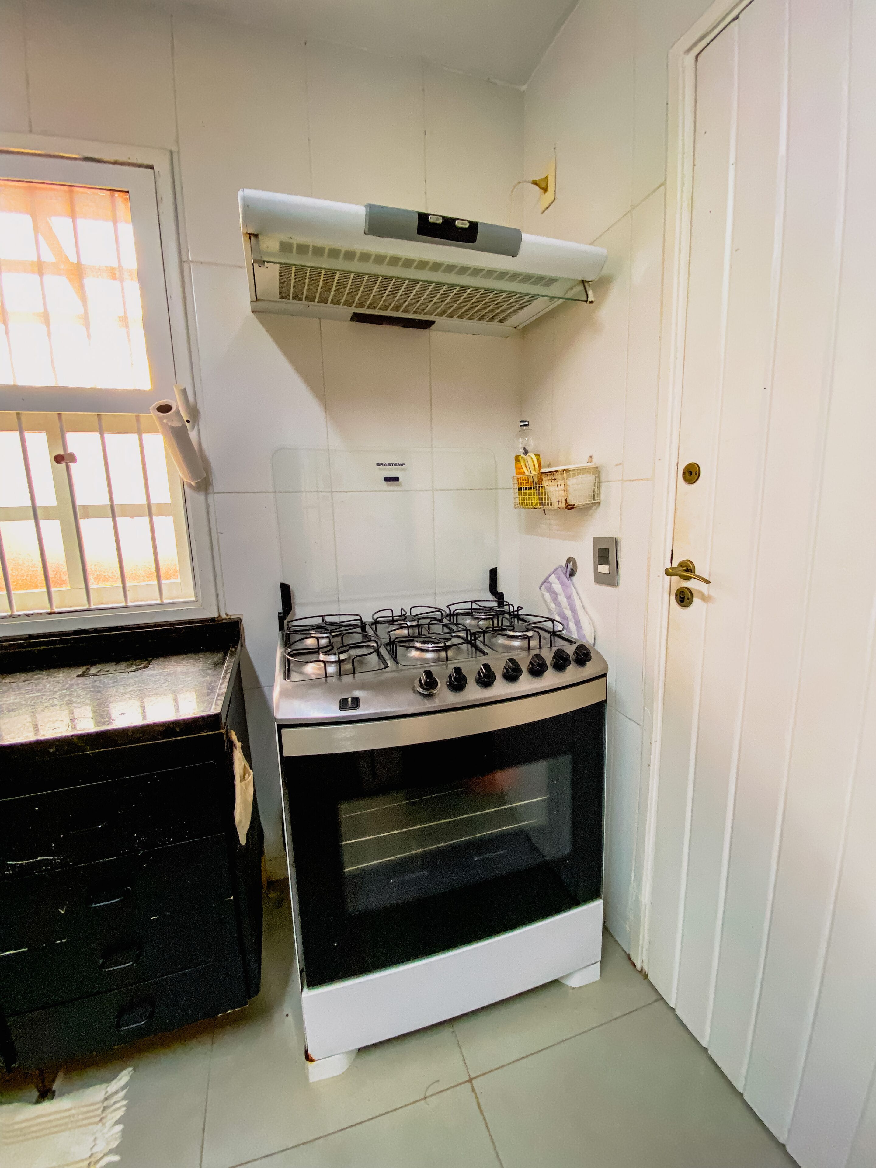 Une cuisine équipée d’une cuisinière à gaz, d’un four, de carreaux blancs, d’une fenêtre avec des barreaux et d’une porte.