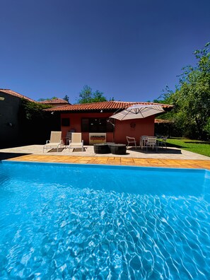 Una piscina con borde de baldosas, mobiliario exterior y un edificio con tejado de tejas rojas.