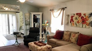 A living room with a sofa, ottoman, and a black chair. There is a ceiling fan, a painting, and a window with curtains. - 435 Flossmoor Court (The Villages)