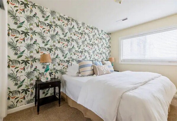 A bedroom with a floral wallpaper, a bed with white bedding, a nightstand, and a window with blinds.